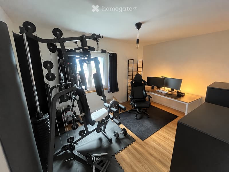 A multi-purpose room with a gym machine on one side and a desk with a computer setup on the other. The room features wooden flooring, a hanging light fixture, and a window with curtains.