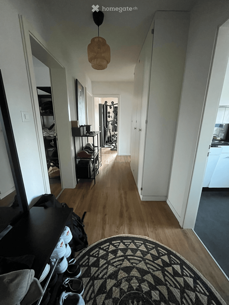 A long, narrow hallway with wooden flooring, white walls, and a hanging woven light fixture. The hallway features several open doors leading to different rooms, including a bedroom, a gym area, and a kitchen. A small side table with items and a shoe rack