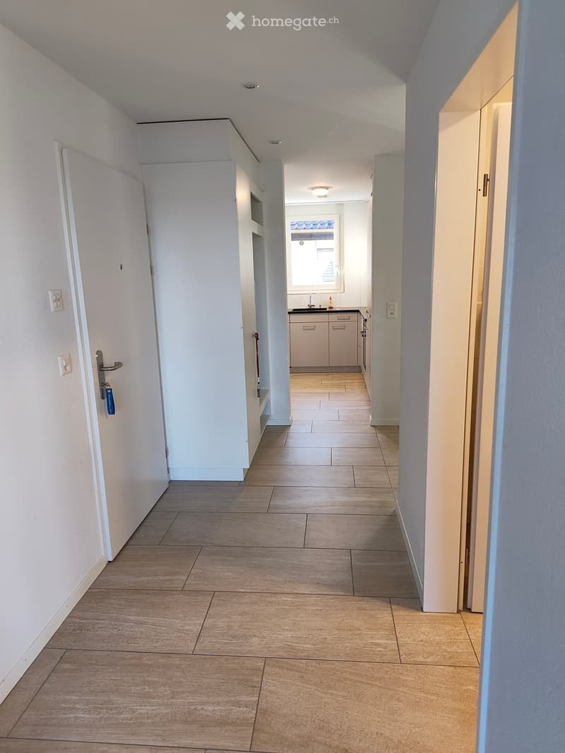 Clean, modern hallway with tiled flooring, white walls, and several doors leading to different rooms, including a visible kitchen area.
