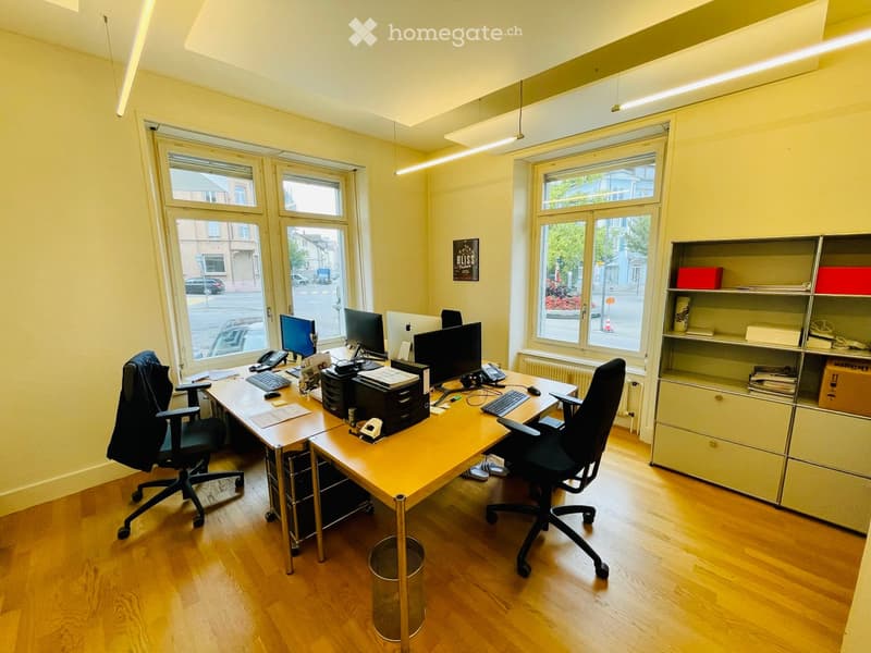 Bright office space with wooden flooring, large windows, a long wooden desk with two office chairs, multiple computer monitors, keyboards, a telephone, and shelves with office supplies.