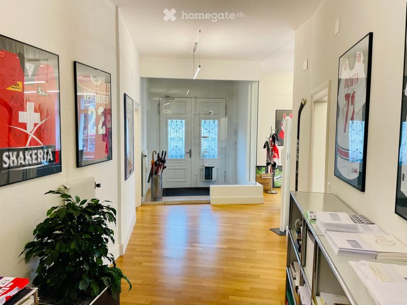 Bright hallway with wooden flooring, white walls, framed artwork, a potted plant, and a glass reception area in the background.