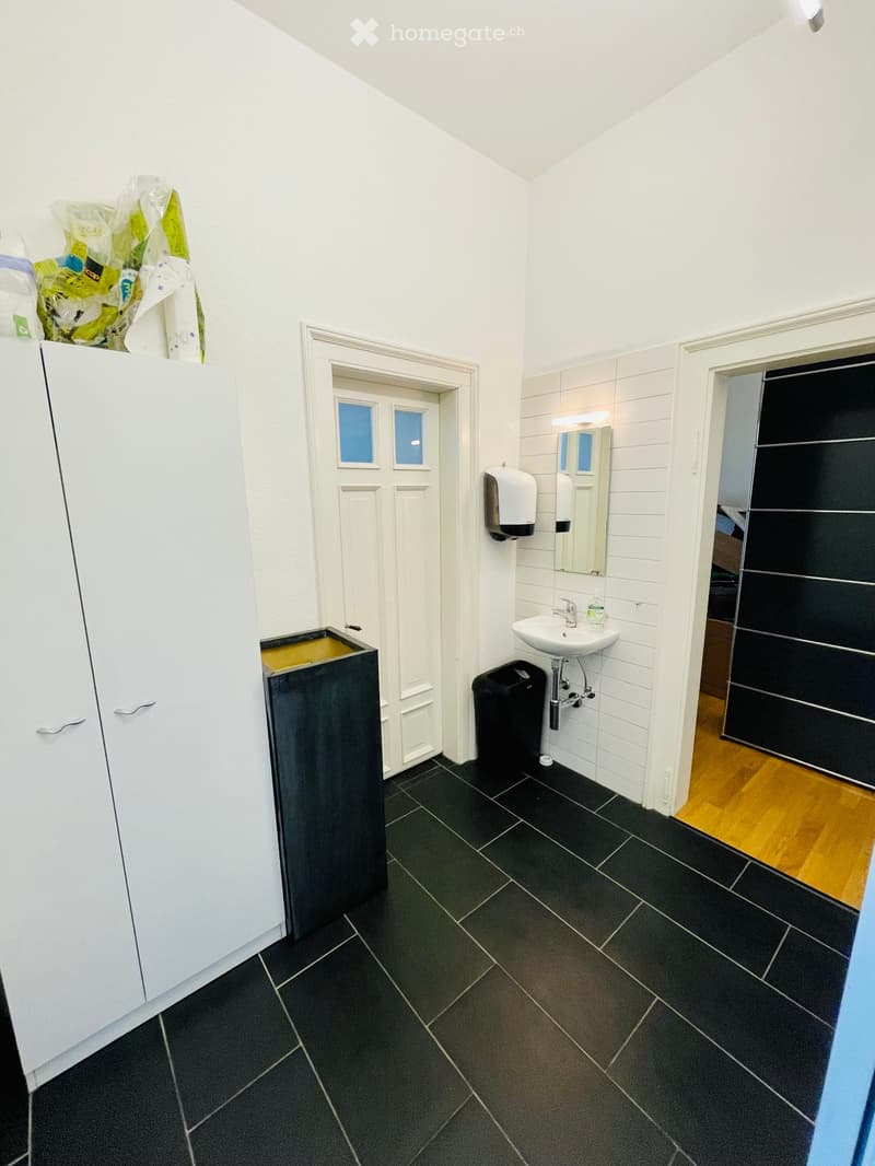 Modern bathroom with black tiled floor, white walls, sink, mirror, cabinet, and trash bin. A door leads to another room with wooden flooring visible.