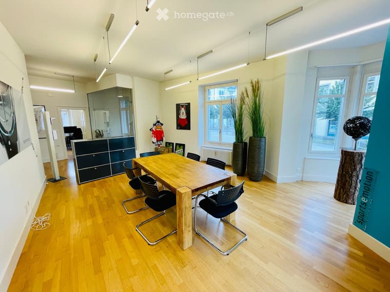 Bright, modern room with wooden flooring, large windows, a wooden table with chairs, a cabinet, and decorative plants.