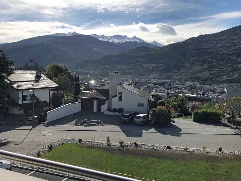 View from a balcony showing a residential area with houses, parked cars, and a scenic mountain backdrop.