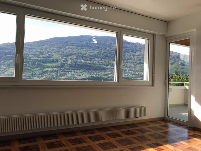Empty room with wooden flooring, large windows offering a scenic mountain view, and a sliding glass door leading to a balcony.