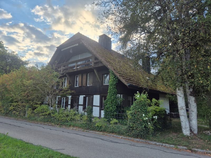 Two-story wooden house with a steep roof, white trim, and a balcony on the upper level, surrounded by trees and greenery.