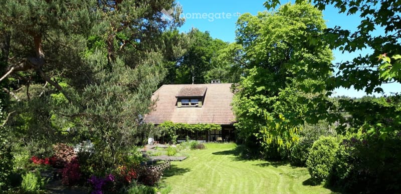 A two-story house with a brown tiled roof, surrounded by a lush garden with trees, shrubs, and colorful flowers. The house has multiple windows and a small porch area.