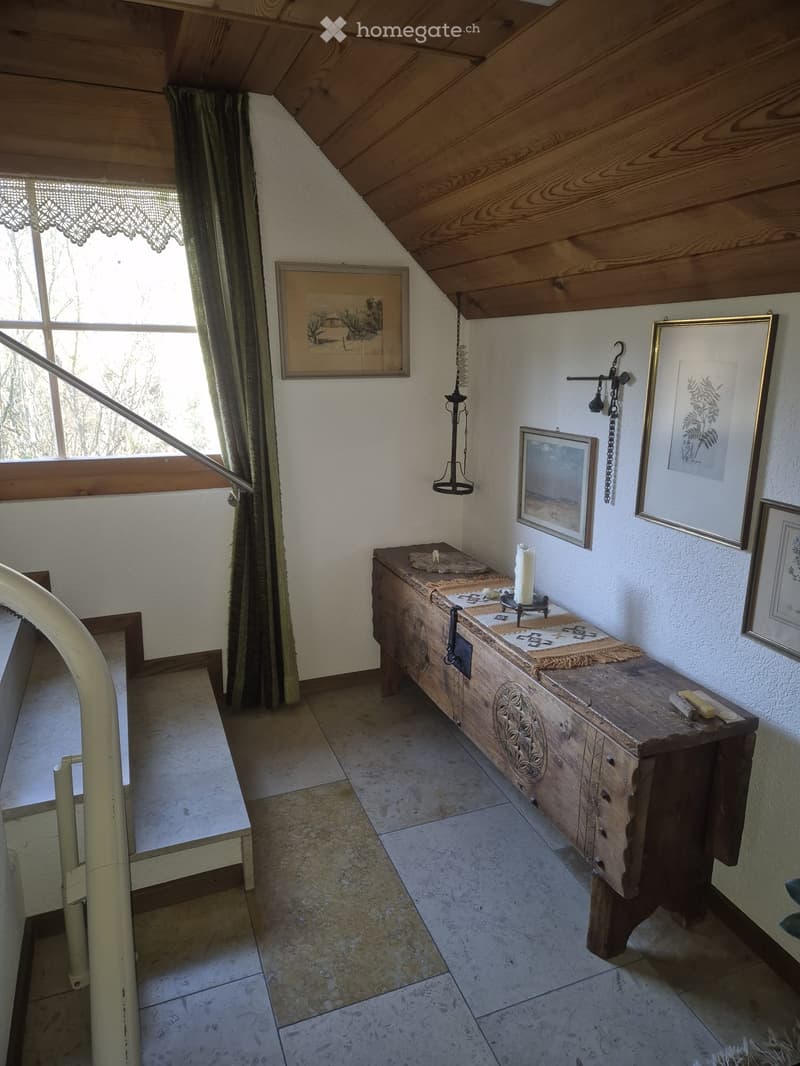 A cozy, wooden-paneled room with a slanted ceiling, featuring a wooden chest with drawers, a candle, picture frames on the wall, and a chair near a window with green curtains.