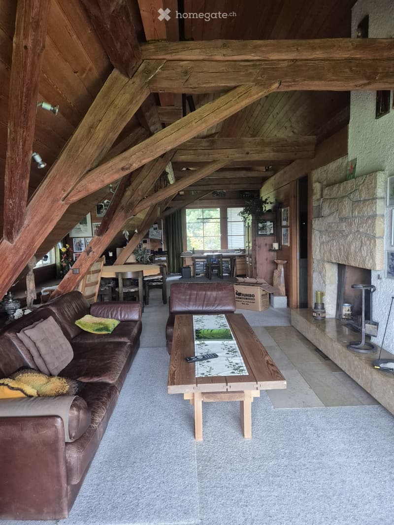 Living room with exposed wooden beams, a brown leather sofa, a wooden coffee table, a fireplace, and a dining area in the background.