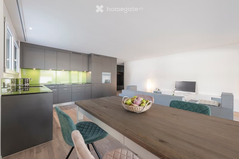 Modern open-plan living and dining area with grey cabinets, green backsplash, wooden dining table, sofa, and chairs.