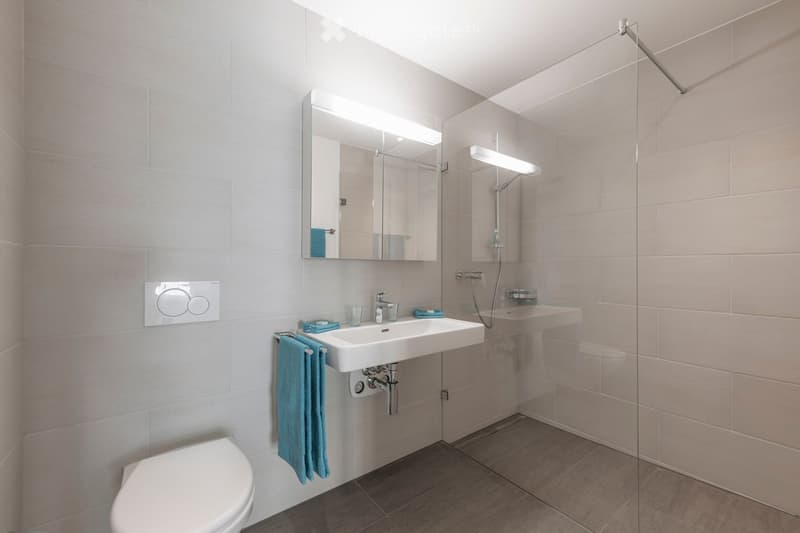 Modern bathroom with a white toilet, sink, mirror, and glass-enclosed shower. Light-colored tiles and a sleek design.