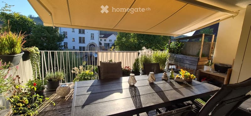 Balcony with a wooden table, chairs, potted plants, and decorative items, featuring a view of a residential area with trees and houses.