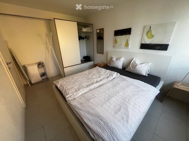 Double bed with 2 pillows, fitted wardrobe with clothes hanging, small side table, wooden floor.