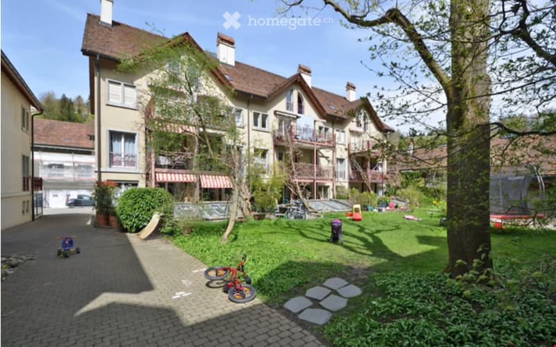 A well-maintained garden area with a paved walkway, green lawn, trees, and a large multi-story building with balconies and awnings in the background.