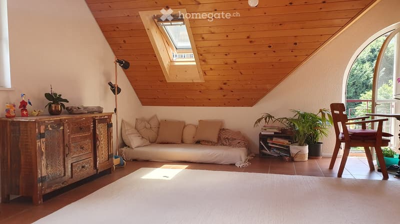 Cozy living room with wooden slanted ceiling, skylight window, sofa, wooden cabinet, chair, and plants.