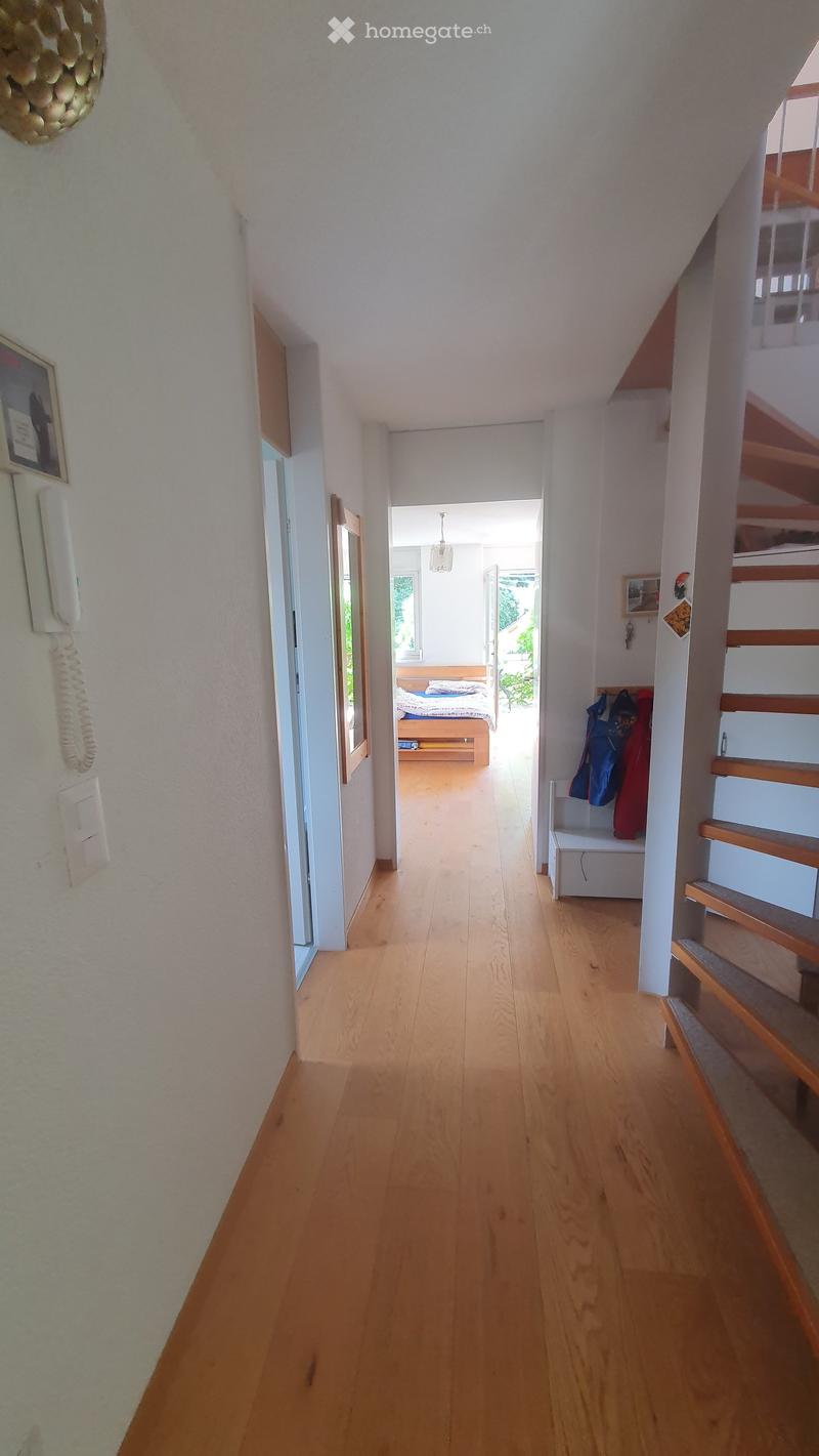 A narrow hallway with wooden flooring, white walls, and a staircase leading upwards. A small bench with coats is visible on the right, and a bedroom is visible at the end of the hallway.