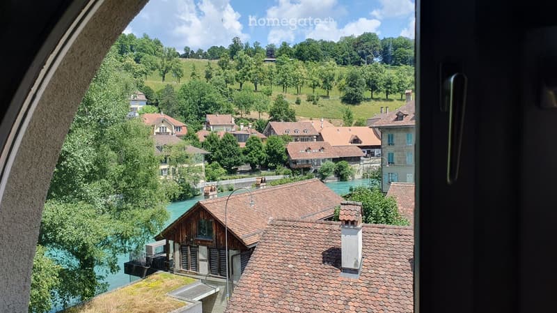 View from a balcony overlooking a picturesque town with red-roofed houses, a river, and lush green hills under a partly cloudy sky.