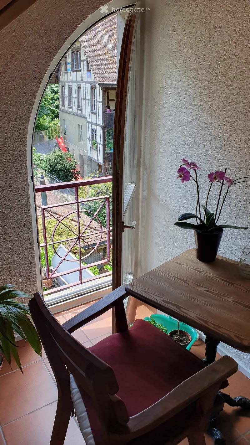 Balcony with a wooden chair, small table, and potted orchid, overlooking a historic building and garden.