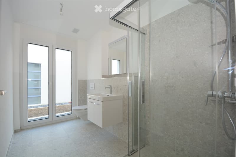 Modern bathroom with a white sink, toilet, mirror, and a glass-enclosed shower. The room features large windows and light-colored tiles.