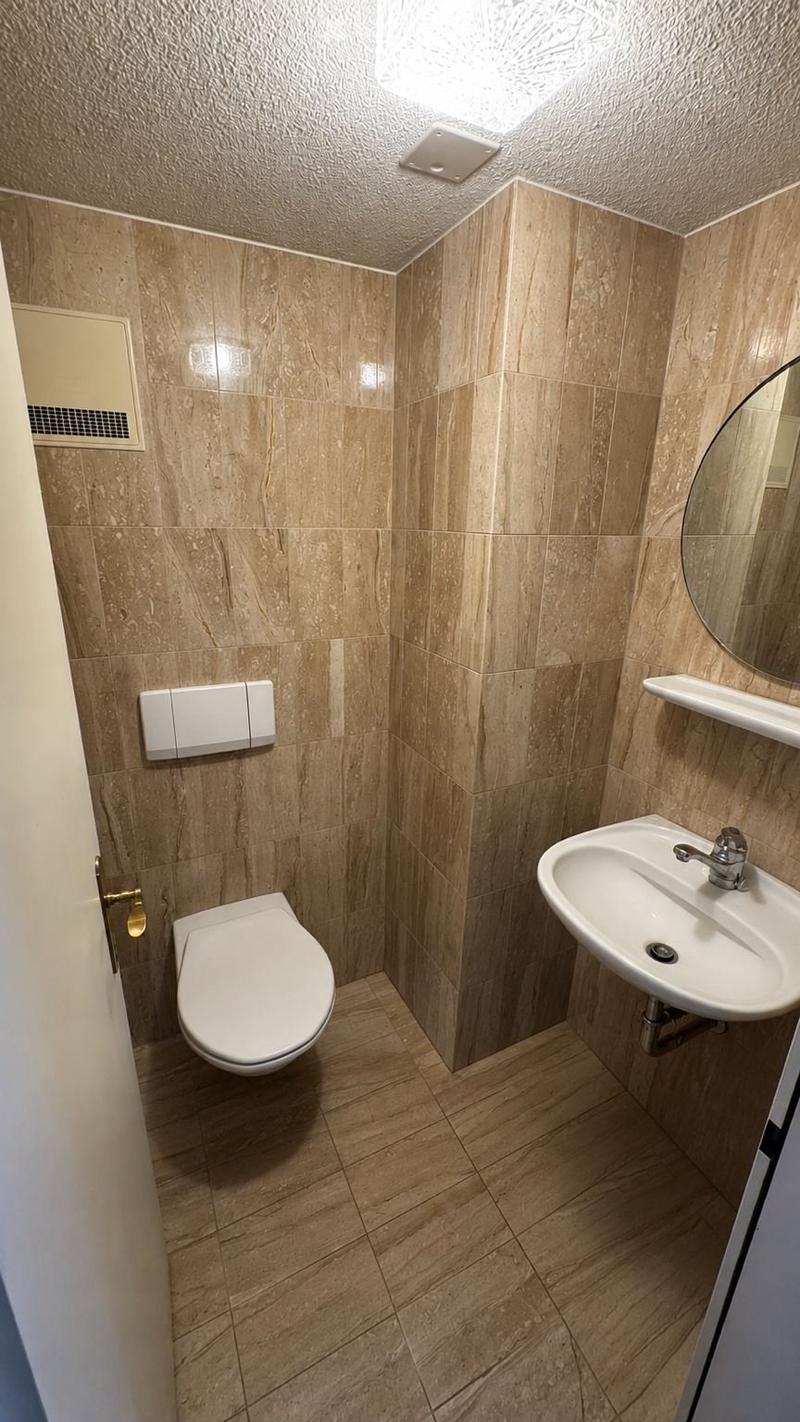 Modern bathroom with beige marble tiles, a toilet, a sink with a chrome faucet, a mirror, and a small shelf.