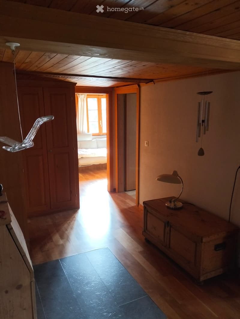A cozy, wooden-floored corridor with a small table and lamp, leading to a bedroom with a sofa and window view.