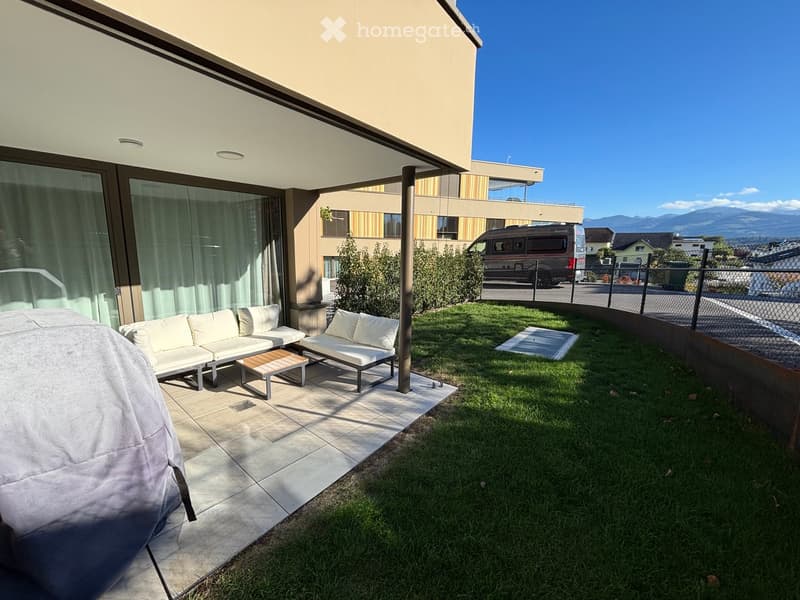 Outdoor patio area with a white sofa, chairs, and a small wooden table, surrounded by a grassy lawn and a modern building in the background.