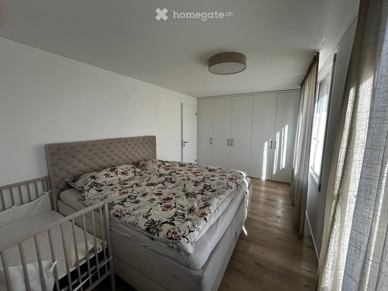 Modern bedroom with a large bed, floral duvet, headboard, and built-in wardrobes. Light wooden flooring and large windows with curtains.