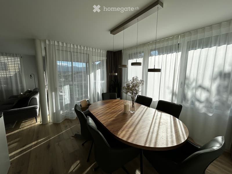 Bright dining area with a round wooden table, six chairs, a candle, a vase with dried flowers, and a sofa visible in the background.