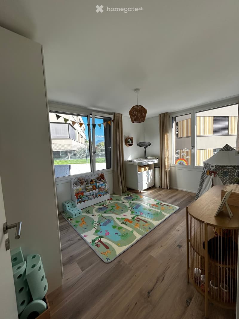 Brightly lit children's room with wooden flooring, large windows, a play mat with animal designs, a small table and chair, a toy tent, a bookshelf, and a storage unit.