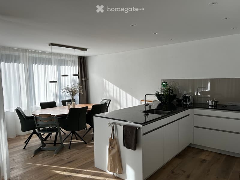 Modern kitchen with white cabinets, dark countertops, a central island, and a dining area with a round wooden table and six chairs. Features include pendant lights, a sink with a modern faucet, and a vase with dried flowers on the dining table.