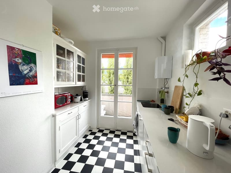 Bright kitchen with white cabinets, black and white checkered floor, and various appliances including a red microwave, blender, and white kettle. Features a large window and a door leading outside.