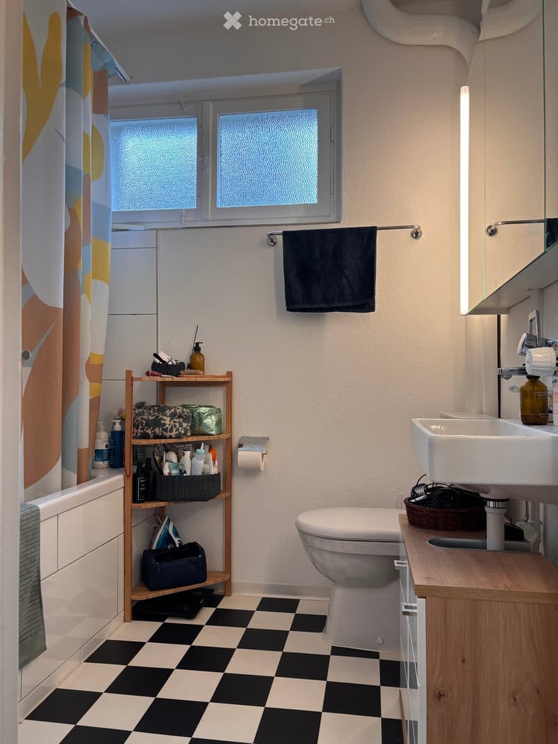 Bathroom with black and white checkered floor, white toilet, sink, bathtub, wooden shelf with various items, towel, and mirror.