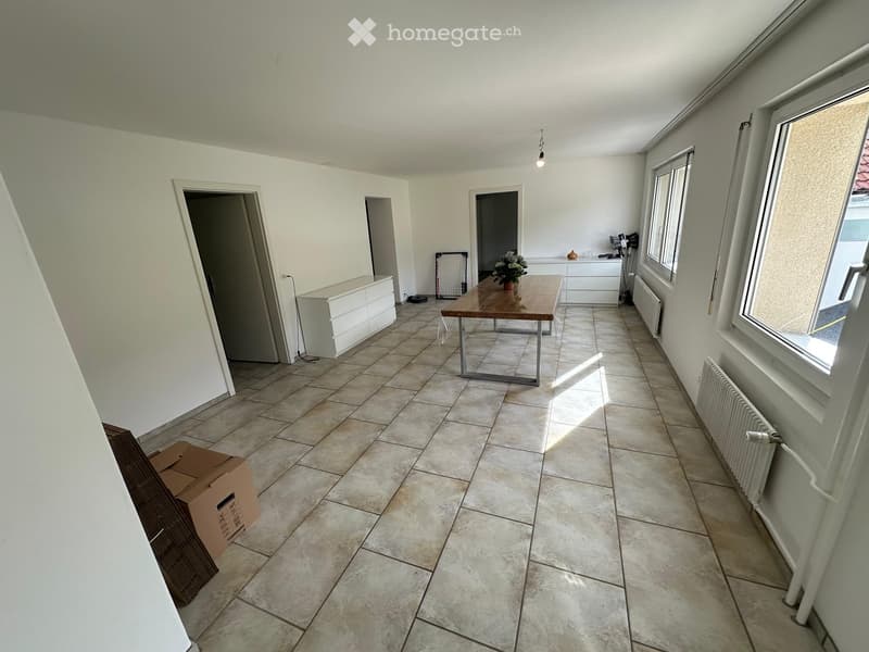 Bright room with tiled floor, white walls, and large windows. Contains a wooden table with a flower vase, white cabinets, radiators, and a cardboard box near the entrance.