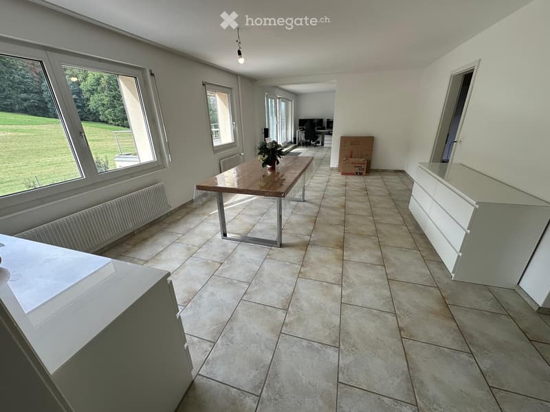 Bright, open room with tiled flooring, large windows, a wooden table with a flower vase, and white cabinets.
