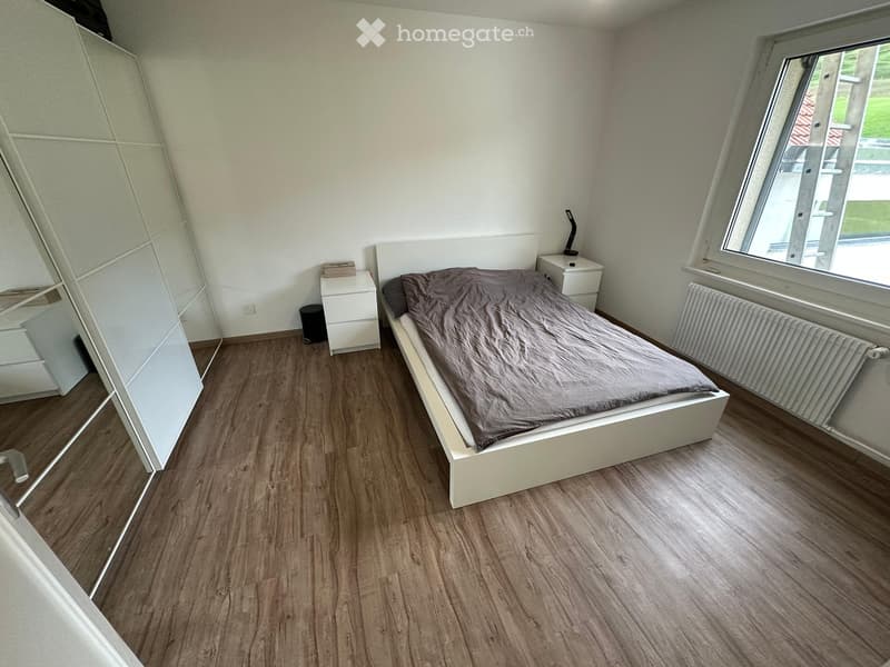 Modern bedroom with wooden flooring, white walls, a bed with grey bedding, a nightstand, a mirror, and a radiator.
