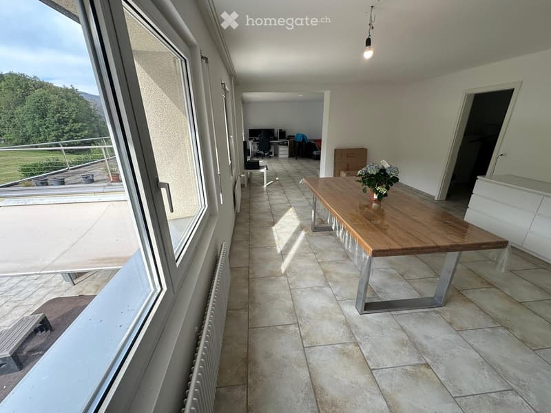 A modern, well-lit hallway with tiled flooring, a wooden table with a flower vase, and large windows offering a scenic view of greenery and mountains.