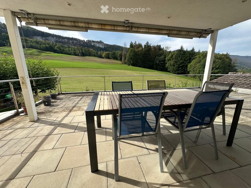 Balcony with a wooden table and blue chairs, overlooking a green landscape with hills and trees.