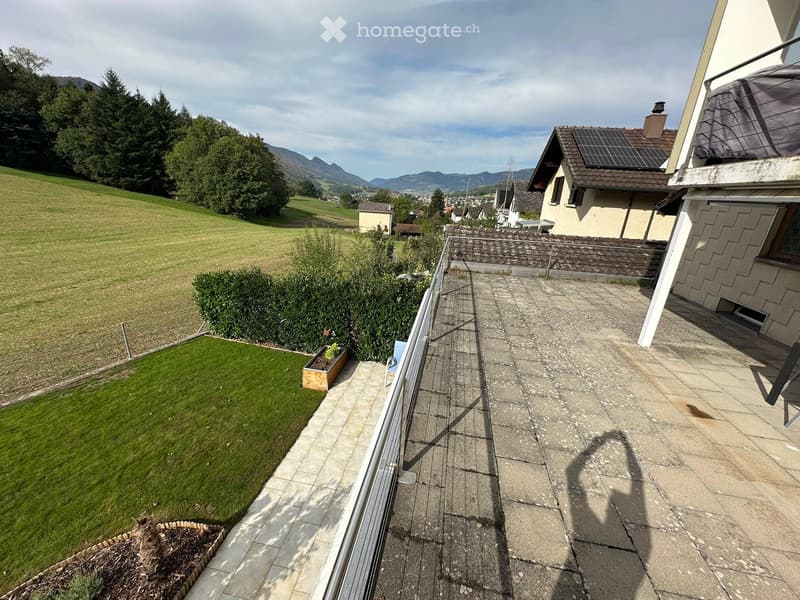 Balcony with stone tiles, metal railing, and a view of a green lawn, trees, and distant mountains.