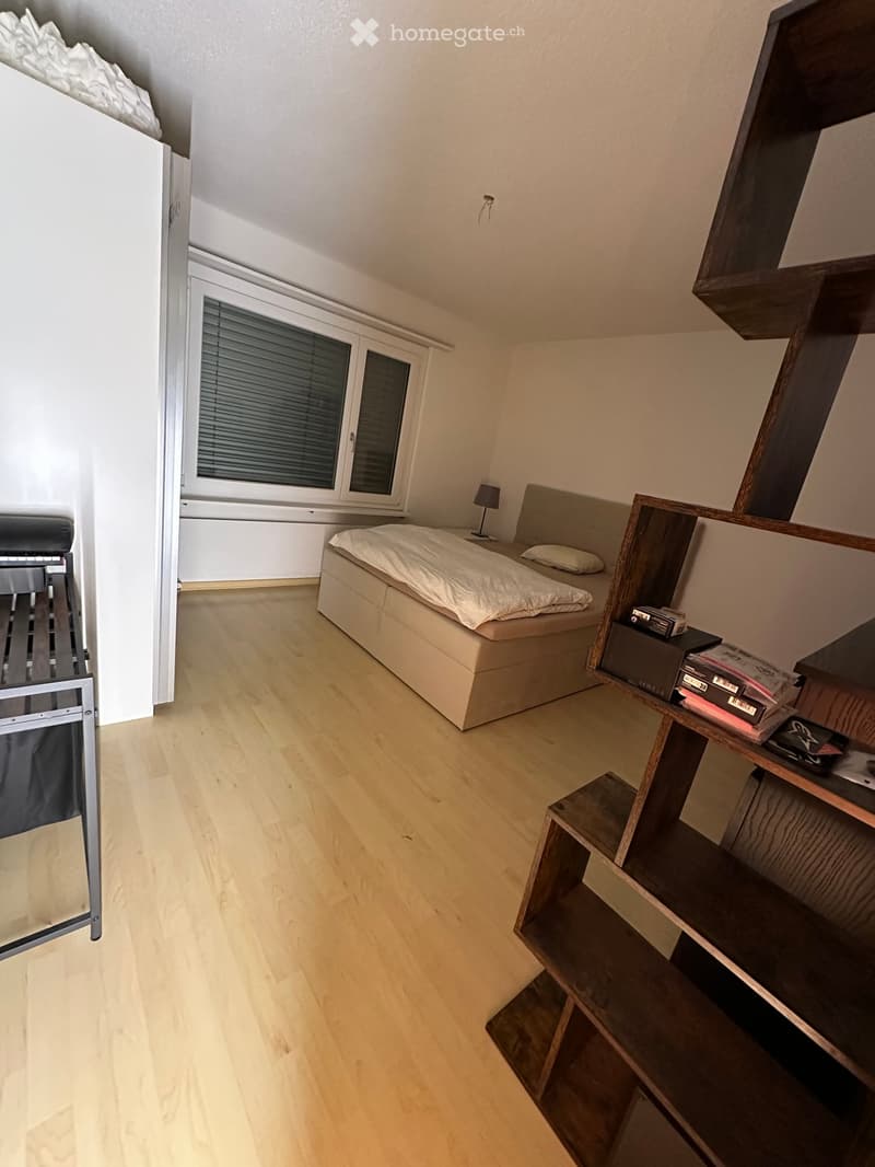 A modern bedroom with a wooden floor, a bed with white bedding, a nightstand with a lamp, and a wooden shelf with books and other items.