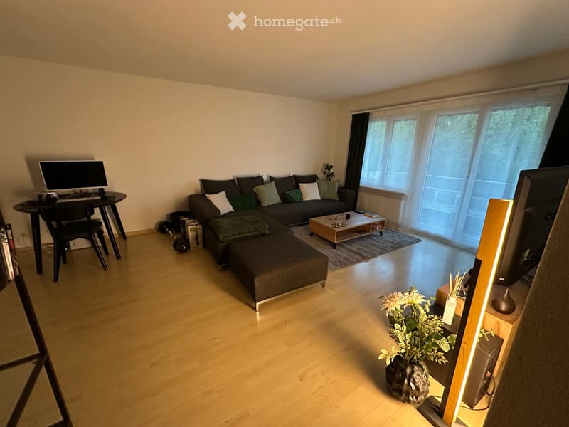 Modern living room with a grey sectional sofa, coffee table, small desk with laptop, and large windows with blinds.