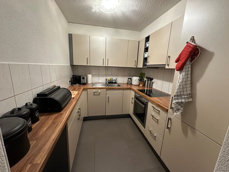 Modern kitchen with light-colored cabinets, wooden countertops, and various appliances including an oven, sink, coffee maker, and toaster. A red oven mitt and a towel are hanging on the wall.