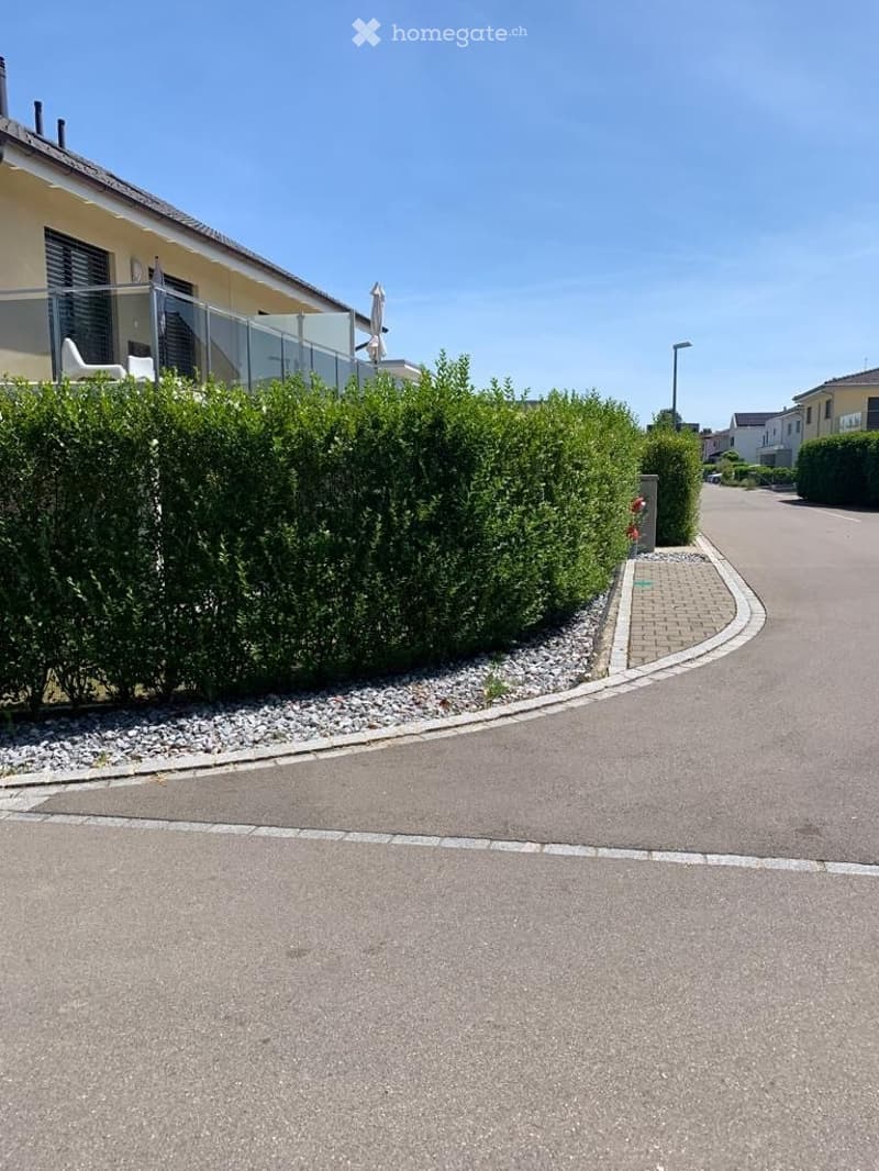 A well-maintained garden area with neatly trimmed hedges, a paved driveway, and a modern house with a balcony in the background.