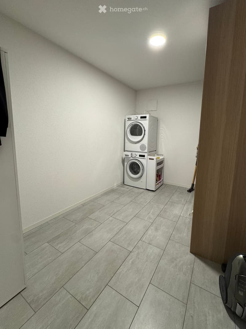 Small, well-lit room with tiled flooring and white walls, featuring a stacked washing machine and dryer unit.