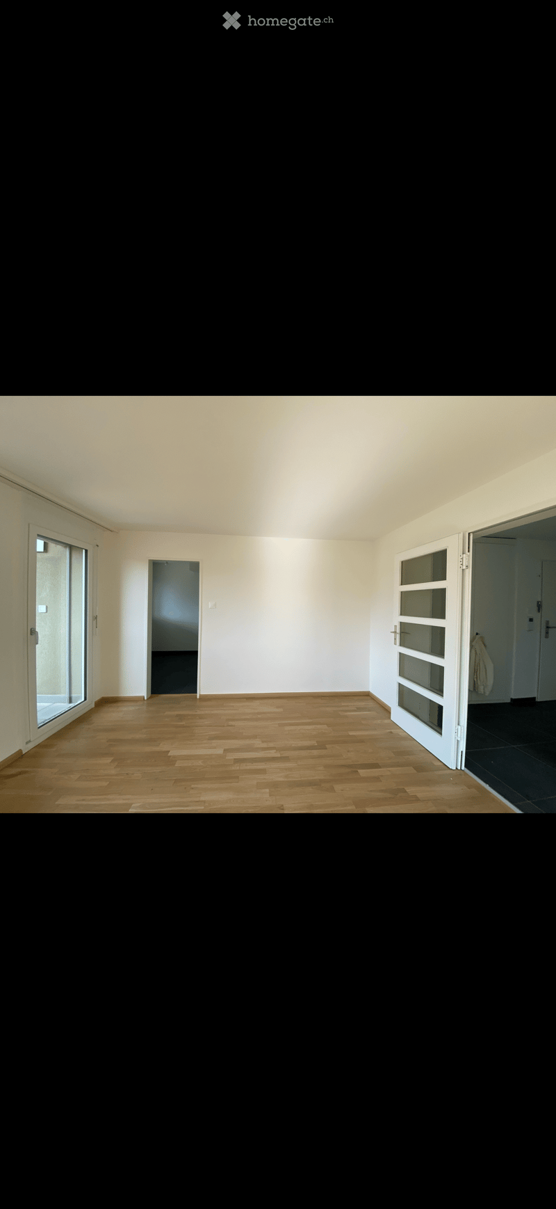 Empty room with wooden flooring, white walls, and large windows, featuring a door leading to another room.