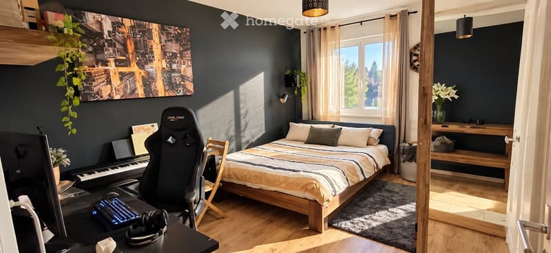 Modern bedroom with a large bed, gaming chair, desk with laptop and keyboard, piano, and decorative plants. Large windows allow natural light to fill the space.