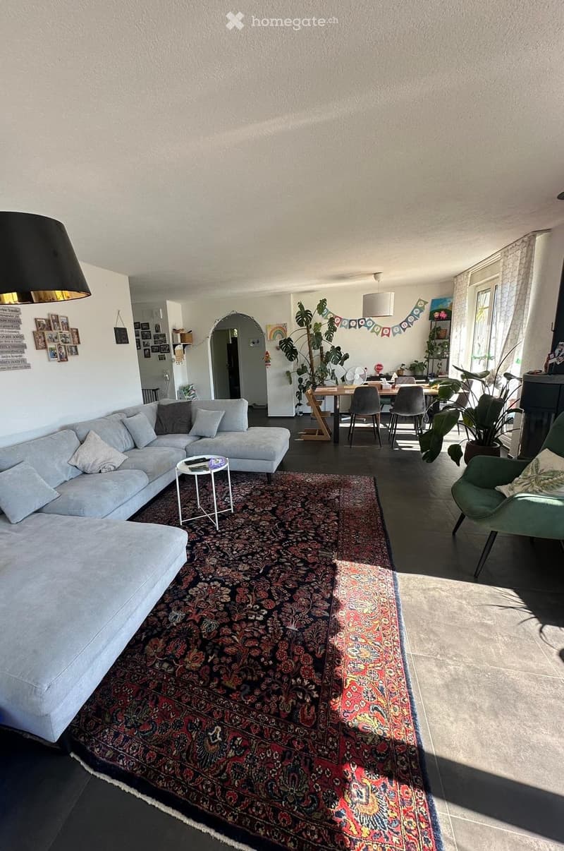 Open-plan living and dining area with a large sectional sofa, coffee table, dining table with chairs, and a green armchair. The space features a patterned rug, large windows, and decorative elements including picture frames and a fan.