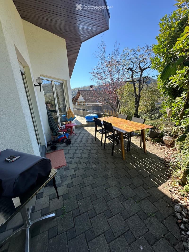 Balcony with a wooden table, chairs, umbrella, grill, and a toy on the floor. The balcony has a tiled surface and overlooks a scenic view with trees and a house in the background.