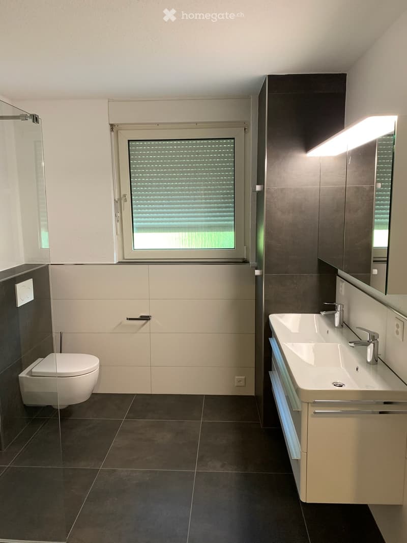 Modern bathroom with a white toilet, double sink, mirrored cabinet, and large window with blinds.