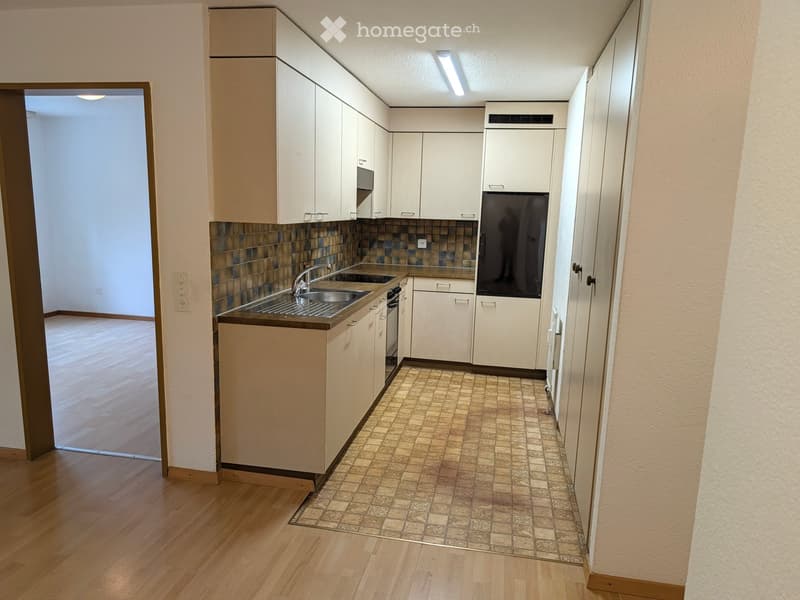 Modern kitchen with white cabinets, stainless steel sink, oven, and refrigerator. The floor is tiled, and the adjacent room has wooden flooring.