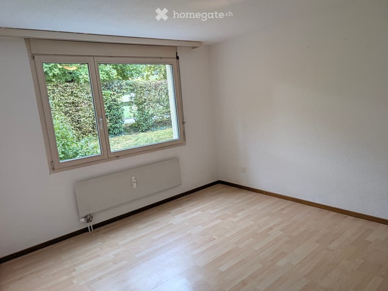 Empty room with light wooden flooring, white walls, a large window with a view of greenery, and a radiator below the window.